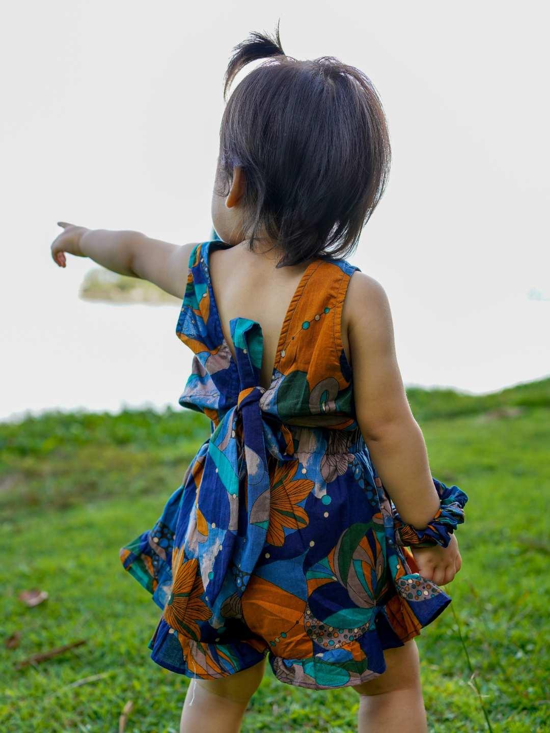 Toddler girl wearing sustainable cotton playsuit in blue and orange mosaic print with open back and bow detail.
