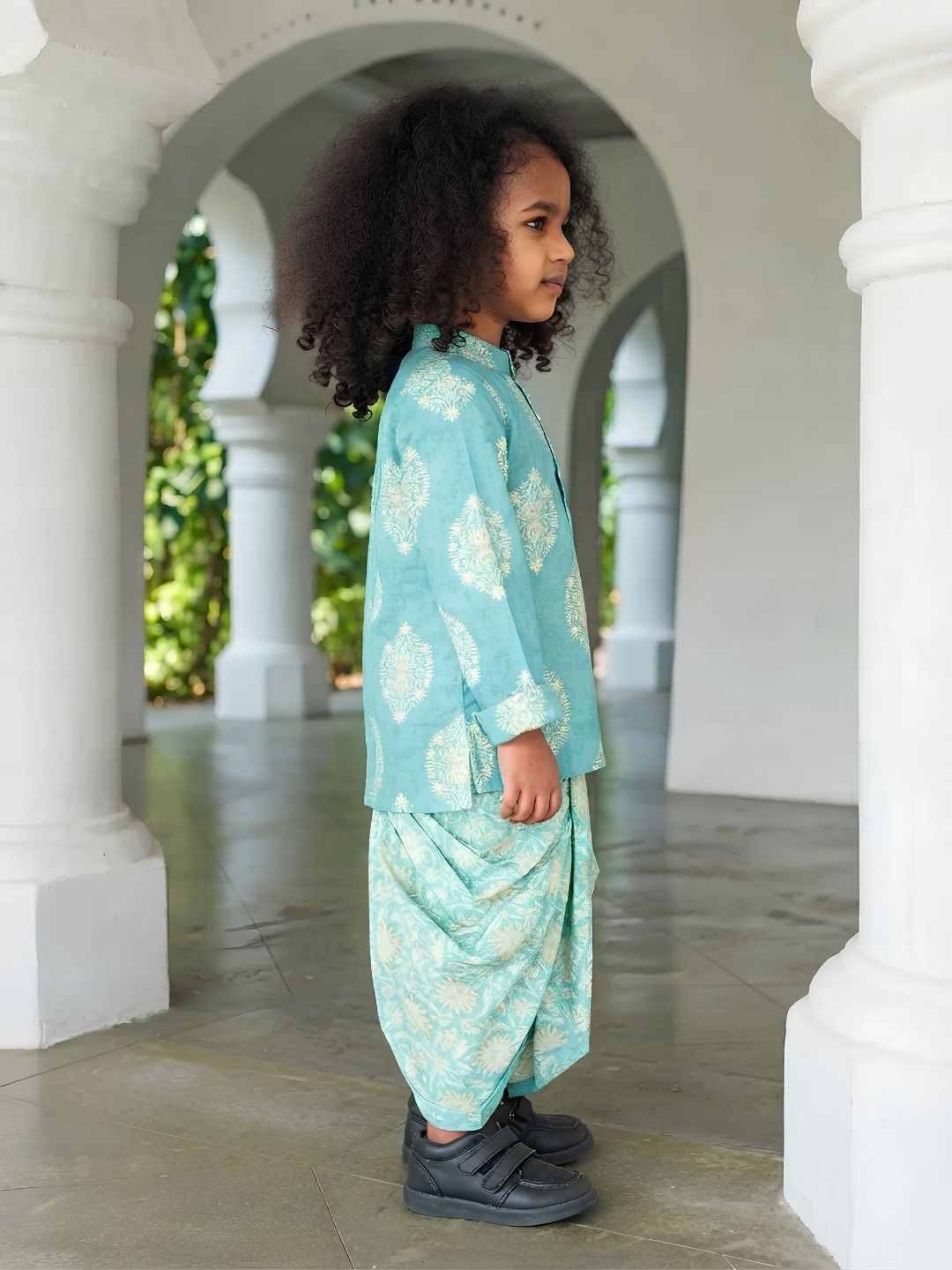 A young child with curly dark hair stands in profile wearing a light turquoise and cream printed traditional outfit with dark gray shoes, posed between white arched columns.