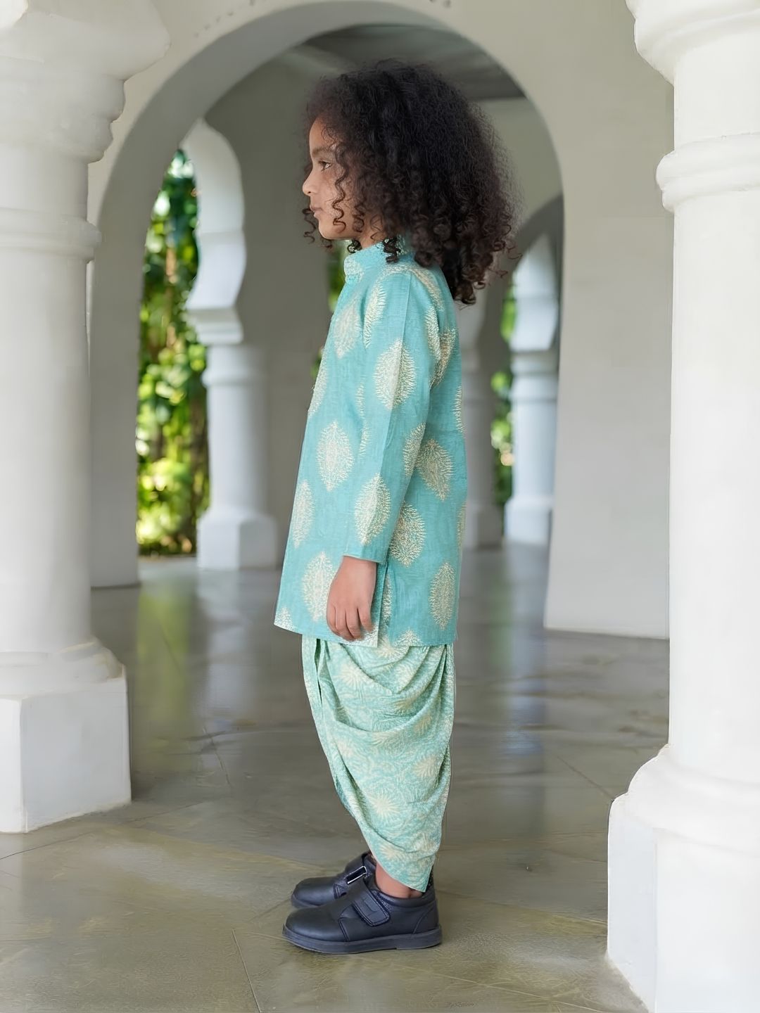 A child model wearing a mint green and white printed kurta and matching harem pants, standing in profile in a white arched corridor.